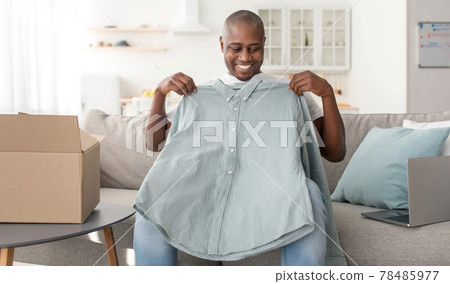 Delivery concept. Happy mature african american man unpacking box and holding new shirt, sitting on sofa 78485977