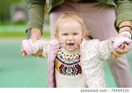 Cute toddler girl having fun on outdoor playground. Young father is teaching his little daughter to walk. First step of baby. Walking for family with little kids. 78494320