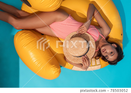 Girl in bathing suit with wicker hat lying on yellow inflatable pool float 78503052
