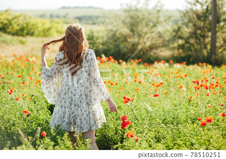 Girl wearing white dress and straw hat walks among red poppy flowers on the sunset. 78510251