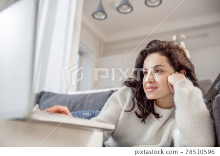 A relaxed woman in white sweater lying on the sofa and watching something online 78510596