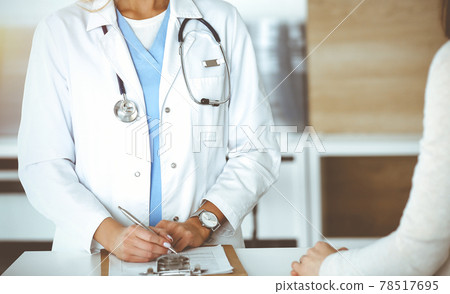 Unknown woman-doctor and patient in sunny hospital. Physician controls medication history records and exam results while using clipboard, close-up Unknown woman-doctor and patient in sunny hospital. Physician controls medication history records and exam results while using clipboard, close-up 78517695
