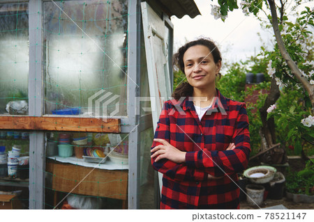 Smiling woman posing to camera with crossed arms on her chest, standing on the background on an old wooden country greenhouse in vegetable garden Smiling woman posing to camera with crossed arms on her chest, standing on the background on an old wooden country greenhouse in vegetable garden 78521147