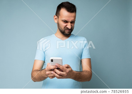 Photo of dissatisfied sad handsome young man with beard wearing everyday blue t-shirt isolated over 78526935