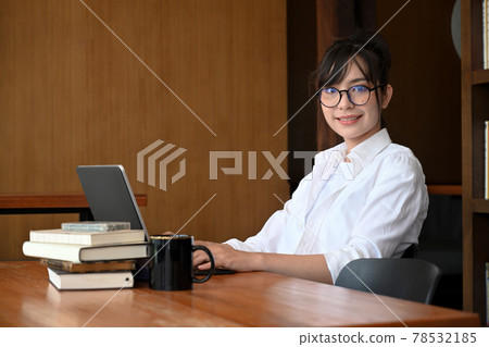 Cheerful female student sitting in library with laptop computer and smiling to camera. Cheerful female student sitting in library with laptop computer and smiling to camera. 78532185