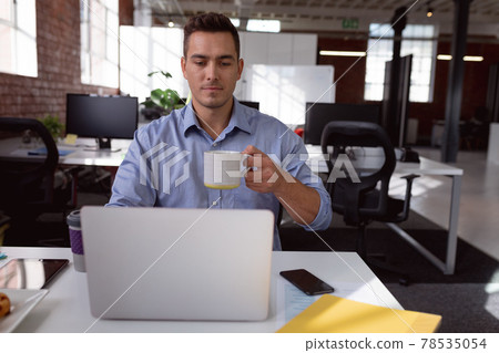 Caucasian businessman sitting at desk using laptop and drinking cup of coffee 78535054