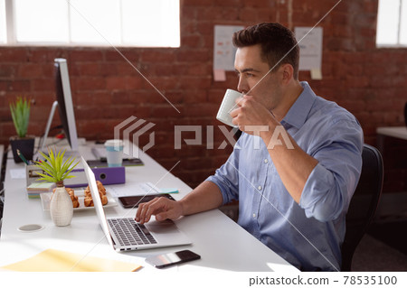 Caucasian businessman sitting at desk in office using laptop and drinking coffee 78535100