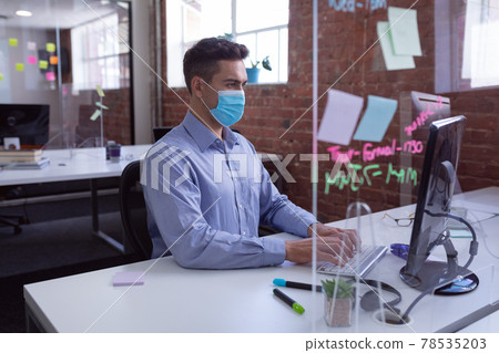 Caucasian businessman in face mask sitting at desk with hygiene screen using computer 78535203