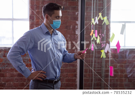 Caucasian businessman wearing face mask brainstorming, reading memo notes on wall standing in office 78535232