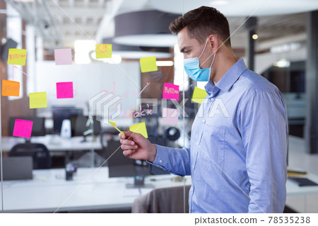 Caucasian businessman wearing face mask brainstorming, reading memo notes on wall standing in office 78535238