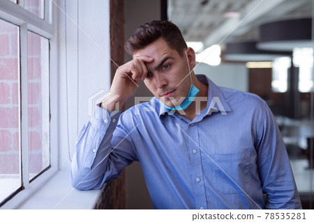 Portrait of concerned caucasian businessman in lowered face mask leaning on windowsill 78535281