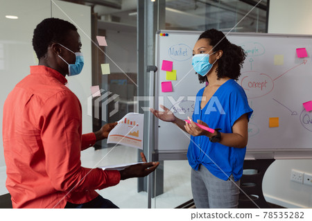Diverse male and female colleague in face masks discussing paperwork in office 78535282