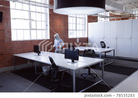 Cleaner wearing ppe suit, glasses and mask disinfecting office workspace 78535283