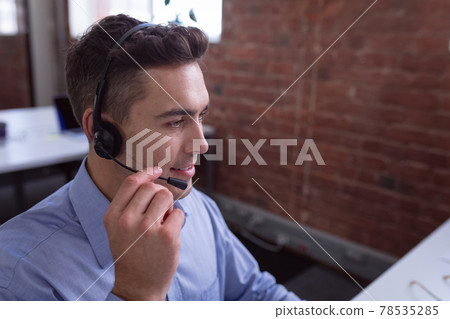 Caucasian businessman wearing phone headset sitting at desk using computer 78535285