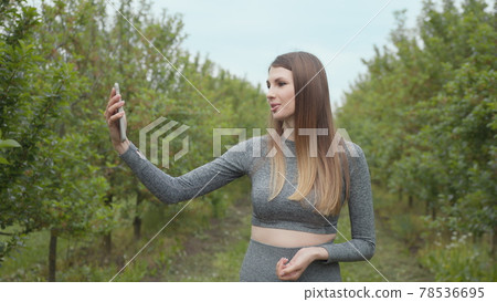 Joyful fitness woman during live stream broadcasting. A modern sports woman takes pictures on the nature smartphone, recording a video blog. Sports video vlog. Video Blog athlete. Girl blogger 78536695