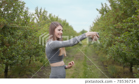 Young Attractive Sports Girl Shooting on a Phone Tree Photographed Against Nature Background. Girl blogger points to the camera nature, the concept of stories about the environment protection of trees 78536714
