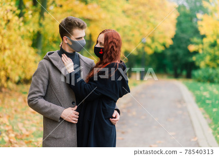 Young red haired woman puts on face mask while walking with young man in autumn park. Young red haired woman puts on face mask while walking with young man in autumn park. 78540313