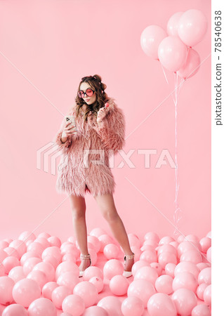 Full length portrait of glamour young woman typing on mobile smartphone over pink balloons background Full length portrait of glamour young woman typing on mobile smartphone over pink balloons background 78540638