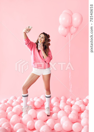 Full length portrait of trendy young woman making selfie photo on smartphone holding milk shake over pink balloons background Full length portrait of trendy young woman making selfie photo on smartphone holding milk shake over pink balloons background 78540658