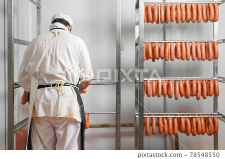 Worker hangs raw sausages on racks in storage room at meat processing factory 78548550