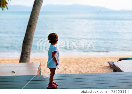 little toddler girl in a white dress looks at the sea while standing on the wooden floor outside. back view little toddler girl in a white dress looks at the sea while standing on the wooden floor outside. back view 78555314