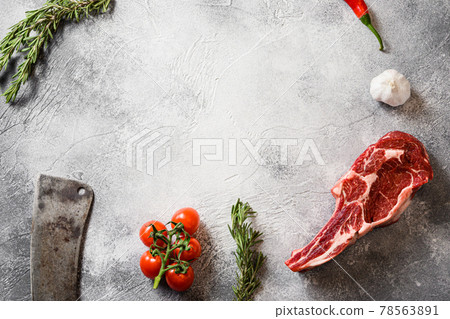 Raw black angus tomahawk steak or Spencer steak cut with the meat american cleaver knife over grey stone table slate cutting top view frame with seasonings, tomatoes, garlic, rosemary. Space for text 78563891