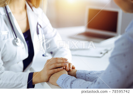 Unknown woman-doctor is holding her patient's hands, discussing current health examination, while they are sitting at the desk in the sunny cabinet in a clinic. Physician with a stethscope at work 78595742