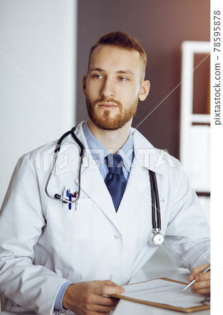 Friendly bearded doctor sitting and writing at clipboard in sunny clinic. Medicine concept Friendly bearded doctor sitting and writing at clipboard in sunny clinic. Medicine concept 78595878