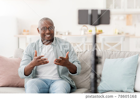 Cheerful mature african american man broadcasting from home, using smartphone and tripod, sitting on sofa at home Cheerful mature african american man broadcasting from home, using smartphone and tripod, sitting on sofa at home 78605168