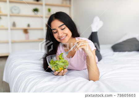 Slimming diet concept. Lovely Indian lady eating tasty vegetable salad on comfy bed at home Slimming diet concept. Lovely Indian lady eating tasty vegetable salad on comfy bed at home 78605603