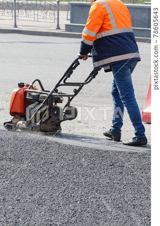 A road worker compacts fresh asphalt with petrol vibratory plate on a sunny day. A road worker compacts fresh asphalt with petrol vibratory plate on a sunny day. 78605643