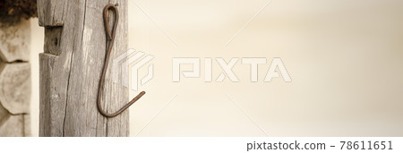 Metal forged hook of brown color on a wooden post of white color. The wall is made of logs and clay. There are cracks and irregularities. 78611651