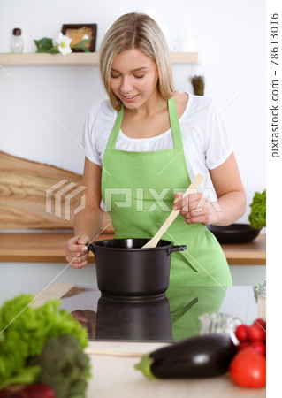 An attractive young blond woman preparing soup by new vegetarian recipe while standing and smiling near the kitchen stove. Cooking and householding concepts An attractive young blond woman preparing soup by new vegetarian recipe while standing and smiling near the kitchen stove. Cooking and householding concepts 78613016