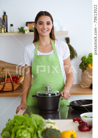 An attractive young dark-haired woman preparing soup by new keto recipe while standing and smiling in the kitchen. Cooking and householding concepts 78613022