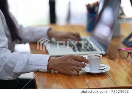 Cropped shot businesswoman holding coffee cup and working with laptop in office. 78625932