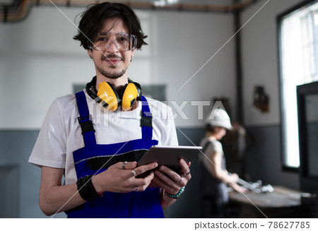 Portrait of young worker with tablet standing indoors in factory looking at camera. Portrait of young worker with tablet standing indoors in factory looking at camera. 78627785