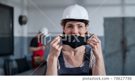 Portrait of woman worker with helmet indoors in factory putting on face mask. Portrait of woman worker with helmet indoors in factory putting on face mask. 78627786