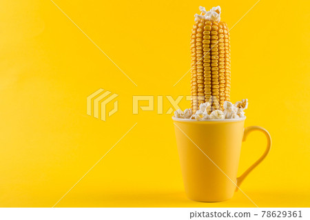 A yellow ear of corn sticking out of a yellow cup, as if growing, popcorn is scattered, on a yellow background, concept 78629361