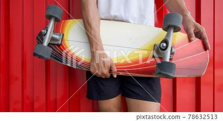 Closeup surfskates board in the hands of teenager in a hip position, in front of red galvanized steel sheet wall. Closeup surfskates board in the hands of teenager in a hip position, in front of red galvanized steel sheet wall. 78632513