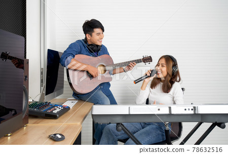 Young asian woman sings while playing an electric keyboard in front of white walls. Her boyfriend played the guitar together. Musicians producing music in professional recording studio. Young asian woman sings while playing an electric keyboard in front of white walls. Her boyfriend played the guitar together. Musicians producing music in professional recording studio. 78632516