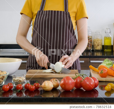 Young chef slices onion into pieces on a wooden chopping board. The kitchen counter full of various kinds of vegetables. Young chef slices onion into pieces on a wooden chopping board. The kitchen counter full of various kinds of vegetables. 78632518