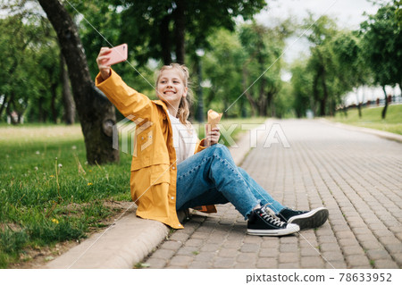 A blonde sitting on the road with a phone and ice cream makes a funny selfie. Child outdoors 78633952