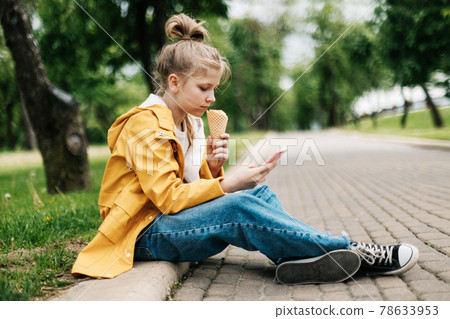 A blond girl sitting on the road with a phone and ice cream communicates in social networks. Child outdoors A blond girl sitting on the road with a phone and ice cream communicates in social networks. Child outdoors 78633953