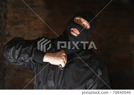 a bandit, a terrorist, a criminal in a black mask holds a knife near his throat, makes a grimace and scares him with murder in a dark room of an abandoned brick building 78640046