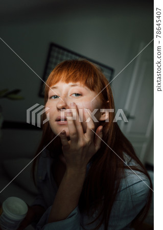 Woman wearing domestic clothes looking at her reflection and applying moisturizer Woman wearing domestic clothes looking at her reflection and applying moisturizer 78645407