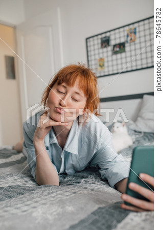 Woman wearing pajamas using mobile phone in bed, while taking a selfie Woman wearing pajamas using mobile phone in bed, while taking a selfie 78645782