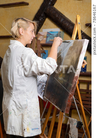 Happy woman paints on canvas using thin brush. Canvas stands on the easel. Artist draws at easel Happy woman paints on canvas using thin brush. Canvas stands on the easel. Artist draws at easel 78646167