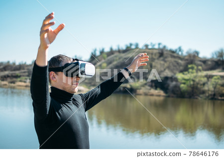 Virtual reality vs real life. Future, Modern technologies. Man in VR virtual reality glasses on the nature background by the river in sun day. Virtual reality vs real life. Future, Modern technologies. Man in VR virtual reality glasses on the nature background by the river in sun day. 78646176