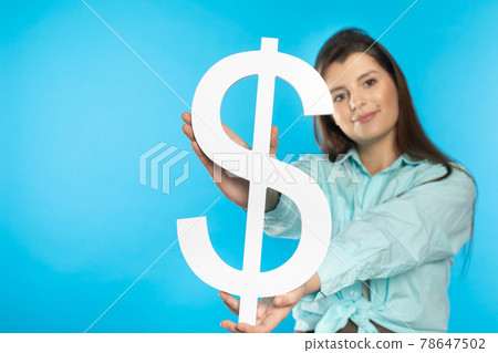 The girl holds a large dollar symbol in her hands. The shape of the US currency. Symbol of finance and cash flow in the economy and the stock market on a blue background. The girl holds a large dollar symbol in her hands. The shape of the US currency. Symbol of finance and cash flow in the economy and the stock market on a blue background. 78647502