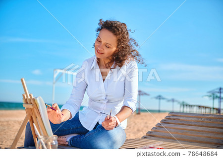 Happy woman artist painting on canvas at the sitting on lonely beach. Art, hobby outdoors . Happy woman artist painting on canvas at the sitting on lonely beach. Art, hobby outdoors . 78648684